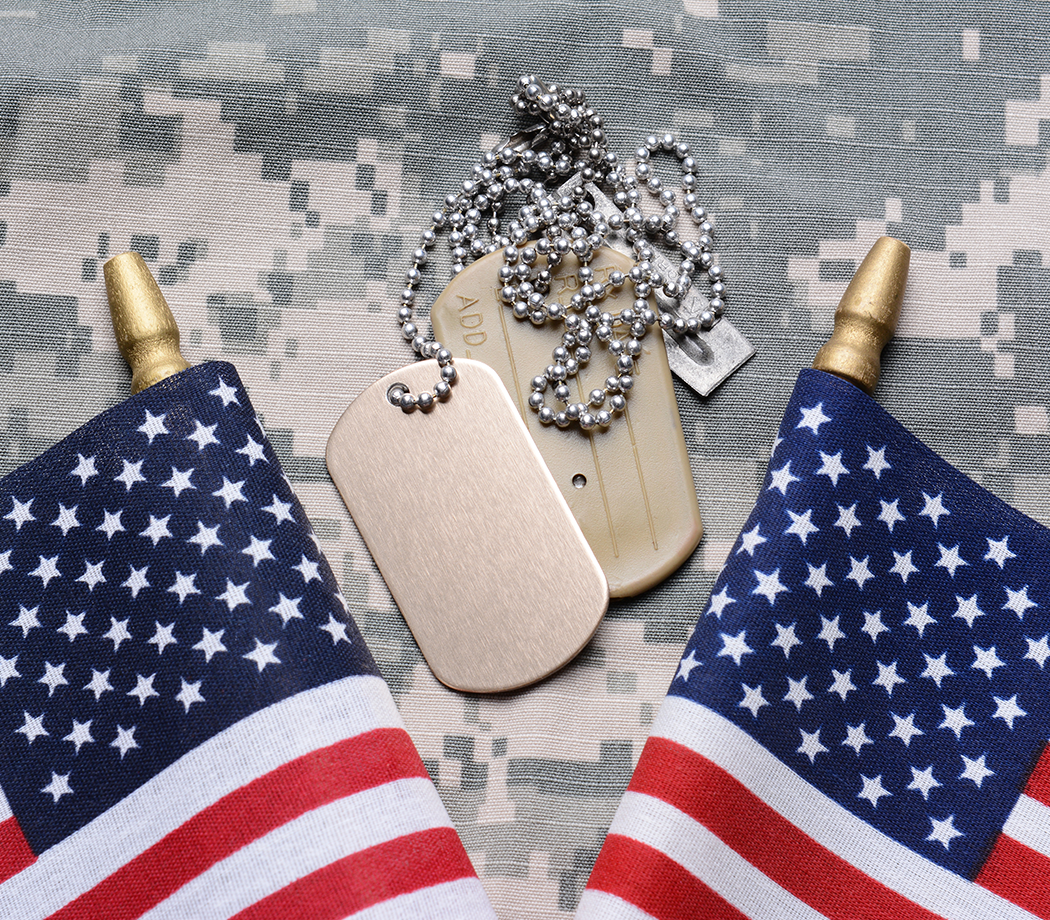 Closeup of two crossed American Flags on camouflage material with dog tags in the middle. The ID tags are blank. Horizontal format filling the frame.