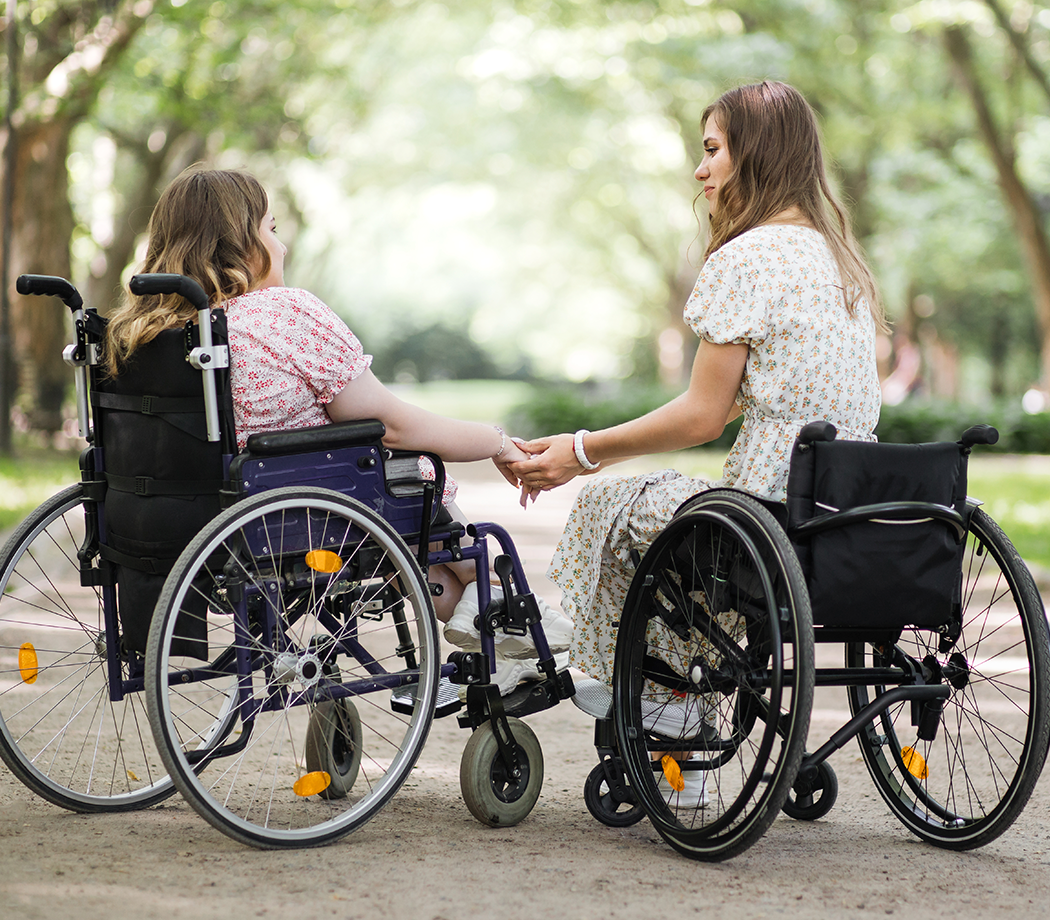 Back view of two young women in summer dressed holding hands while sitting in wheelchairs among green park.