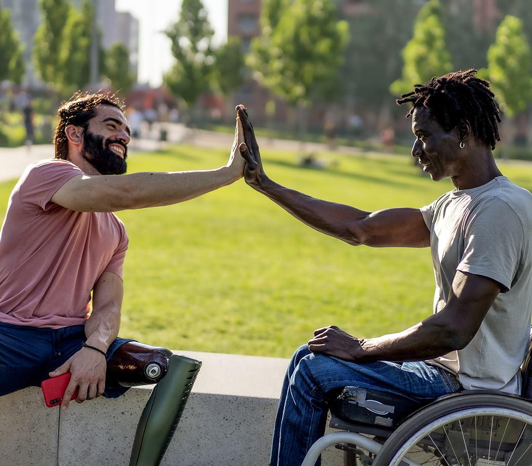Two disabled friends, one Hispanic with an amputated leg prosthesis, the other African in a wheelchair high-fives as a sign of friendship.
