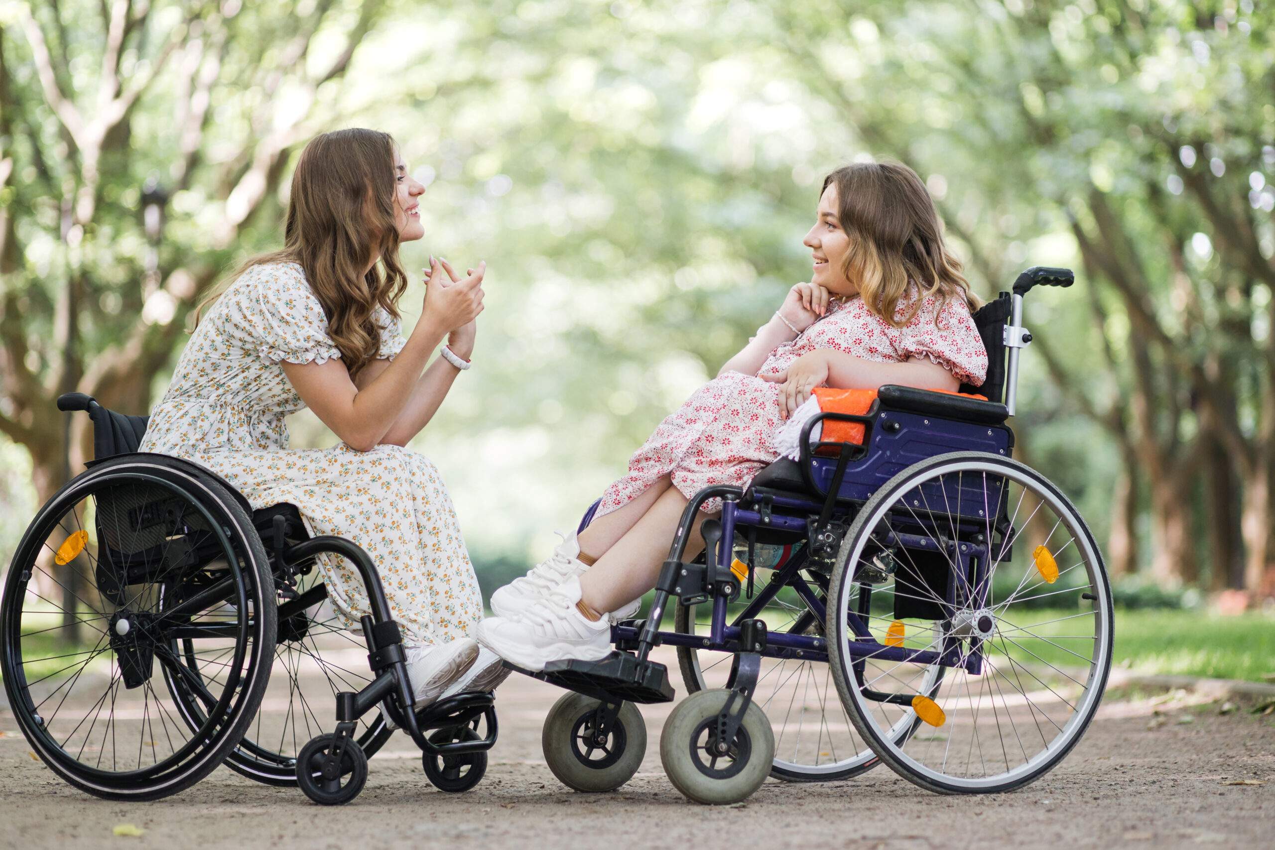 Side view of two happy women in wheelchairs talking and smiling outdoors. Background of green summer park.
