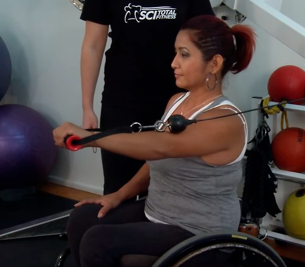 Woman in a wheelchair doing shoulder exercises in a gym setting.