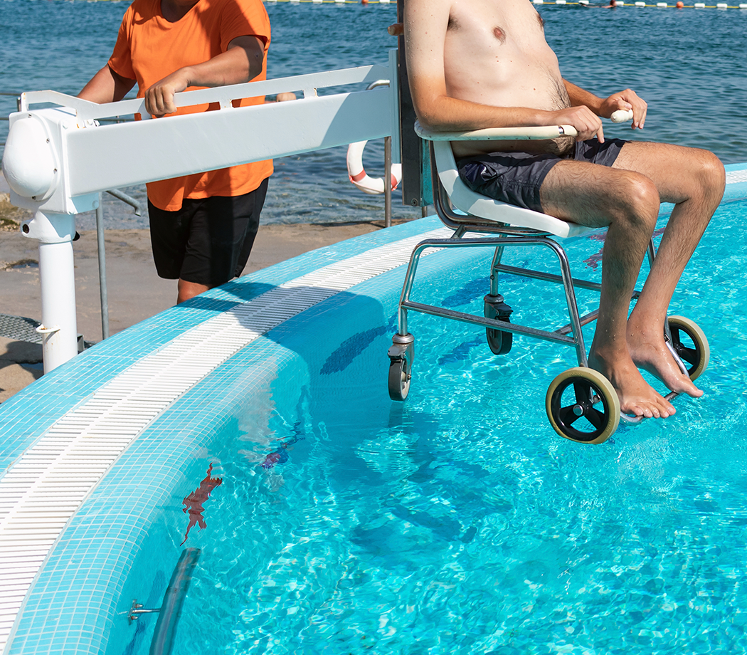 Disabled man on pool wheelchair lift and transferred into water by an assistant.