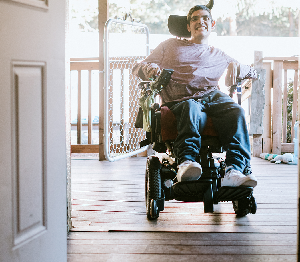 A cheerful young adult man with cerebral palsy drives up a wheelchair ramp to enter his home. Horizontal image with copy space.