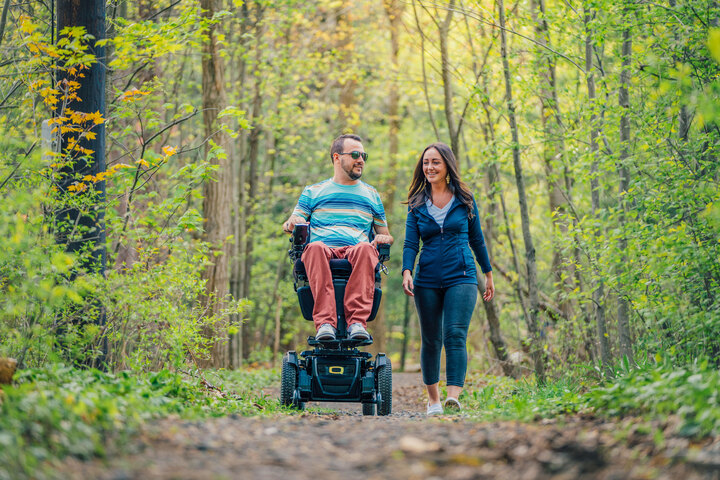 Photo of a man in a power wheelchair and a woman walking on a wooded path.