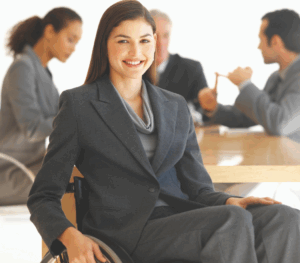 A professional in a wheelchair smiles at the camera, with three colleagues having a discussion in the background.